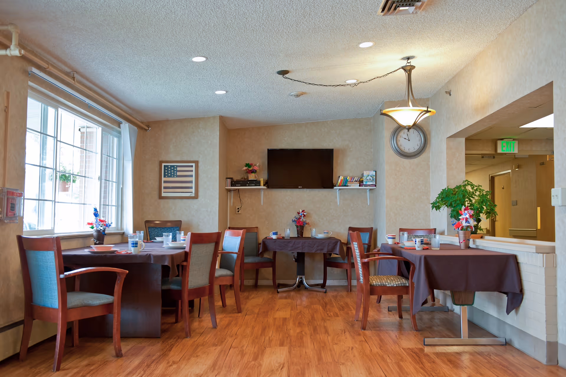 A dining area in a senior living facility with several tables covered in brown tablecloths, each set with cups, plates, and small decorative flower arrangements. The room has wooden flooring, beige walls, a large window letting in natural light, a wall-mounted TV, a clock, and a framed American flag on the wall.