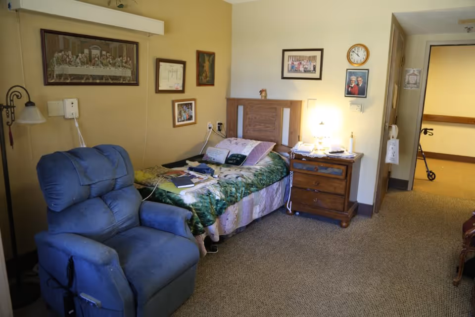 A cozy bedroom in a senior living facility featuring a single bed with a floral quilt, a wooden headboard, and a nightstand with a lamp and various items. A blue recliner chair is positioned next to the bed. The walls are decorated with framed pictures and a clock. A hallway with a walker is visible through an open door.