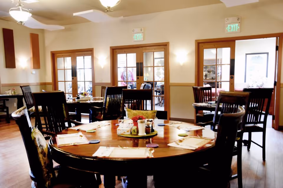 A dining room in a senior living facility with round wooden tables set with placemats, utensils, and condiments. Several wooden chairs surround the tables. The room has wooden floors, cream-colored walls with wooden trim, and multiple glass-paned doors leading to other rooms. Ceiling lights and wall sconces provide illumination.
