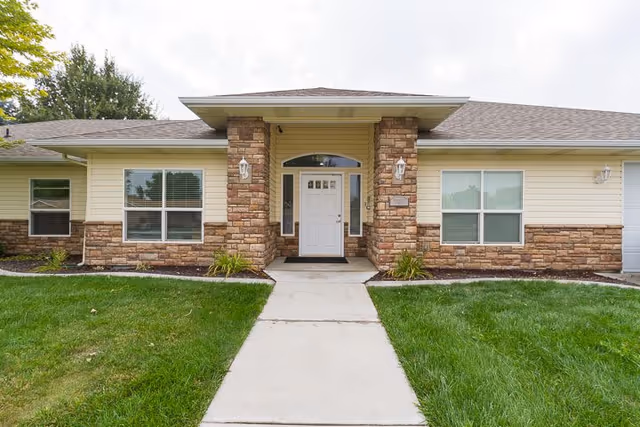 Front exterior view of a single-story building with beige siding and stone accents. The entrance has a white door with sidelights and a small overhang roof. There are two windows on either side of the door and a concrete walkway leading up to the entrance, surrounded by green grass and small plants.