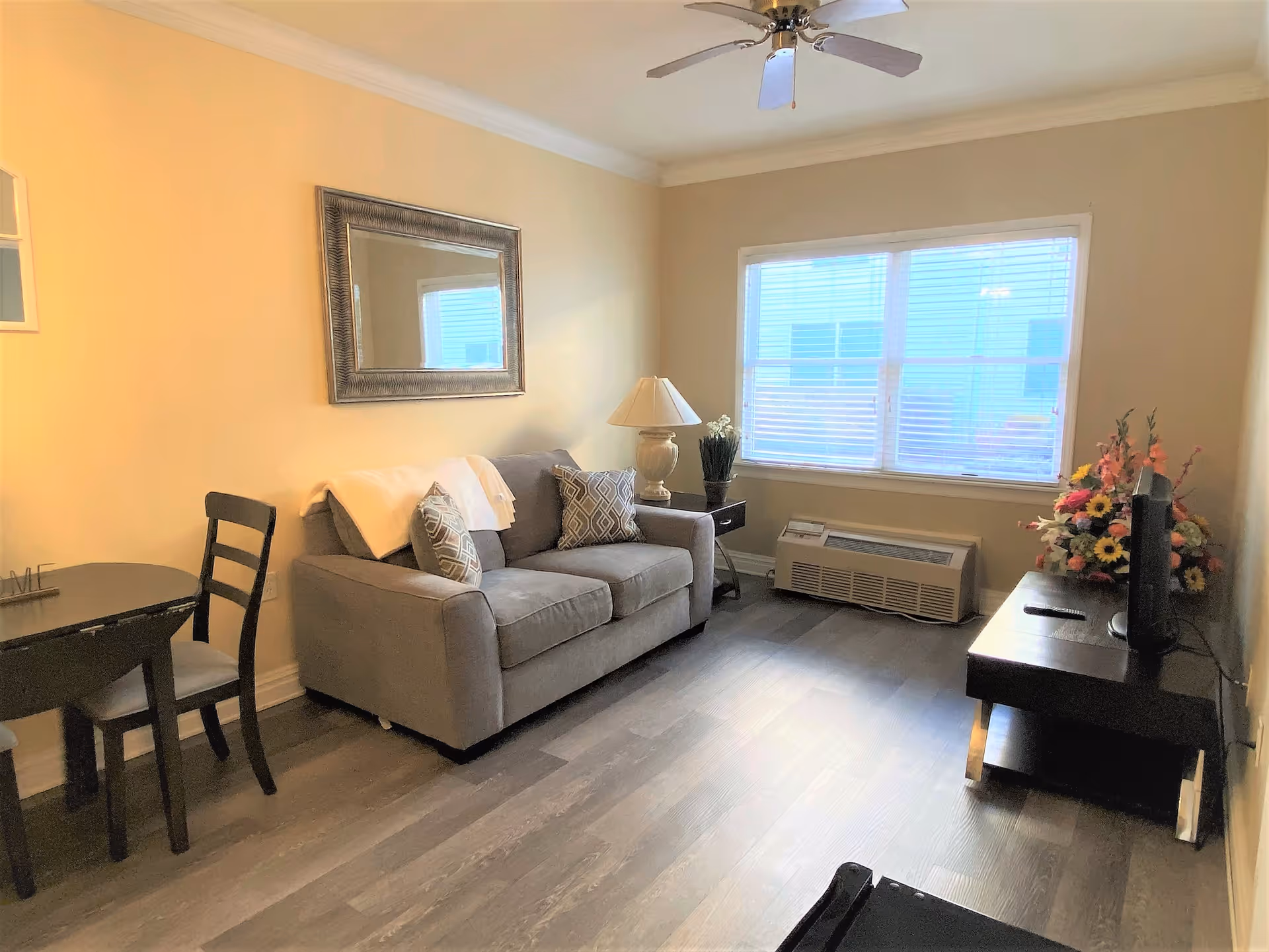 A cozy living room with a gray sofa adorned with patterned pillows and a white throw blanket. A large mirror hangs on the beige wall above the sofa. Next to the sofa is a small side table with a lamp and a plant. A window with blinds lets in natural light. Across from the sofa is a dark wooden TV stand with a flat-screen TV and a colorful flower arrangement. There is also a small dining table with two chairs on the left side of the room. The floor is covered with wood-style flooring and a ceiling fan is mounted above.