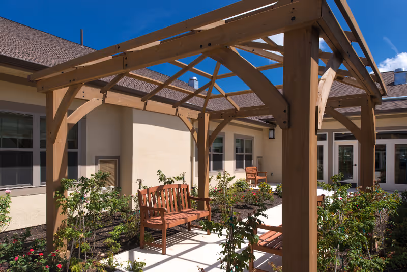 Outdoor courtyard area with wooden pergola, wooden benches, and surrounding garden beds with plants and flowers. The courtyard is enclosed by a beige building with windows and doors under a clear blue sky.