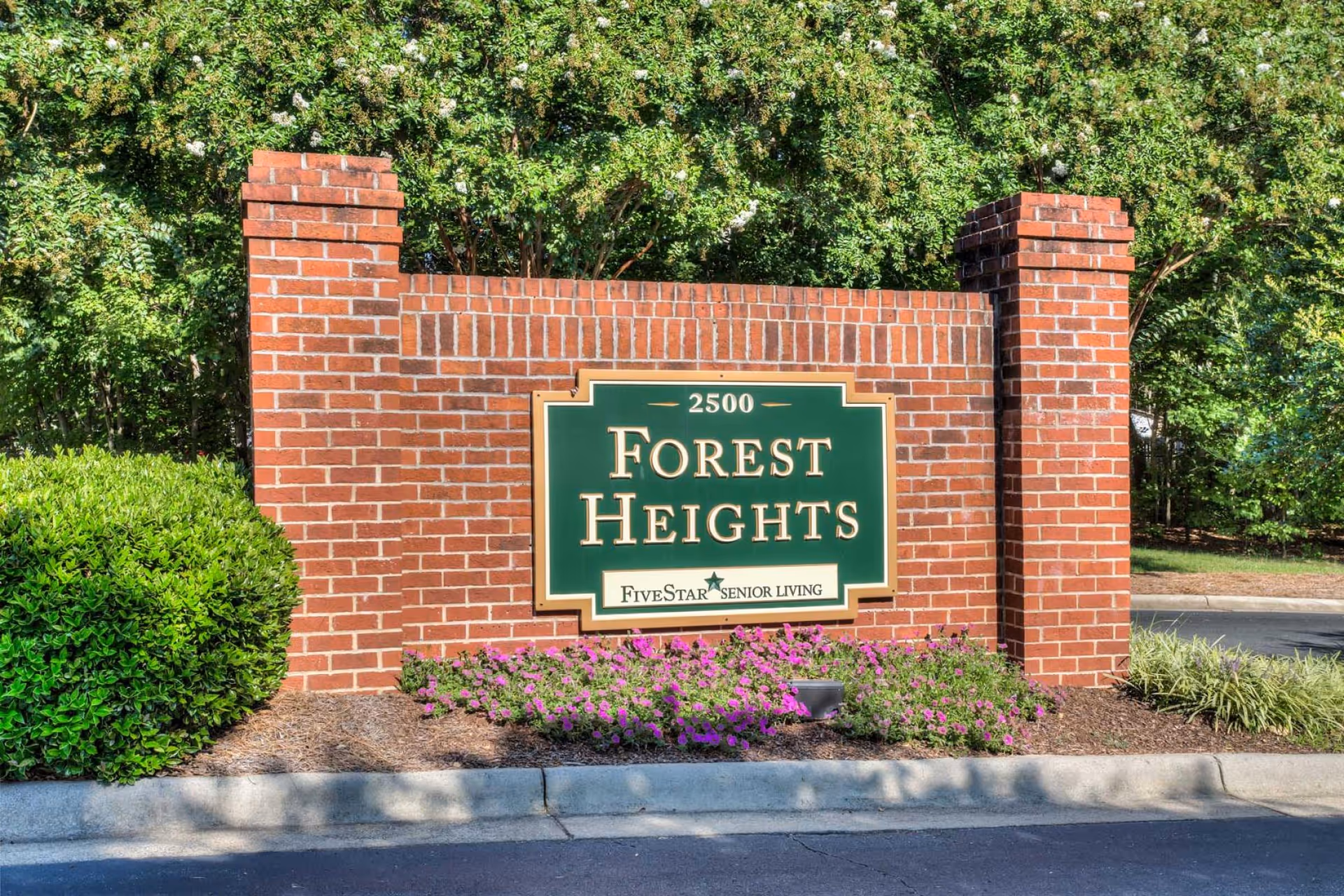 A brick signpost with a green and gold sign that reads '2500 Forest Heights FiveStar Senior Living' surrounded by green bushes and purple flowers, with trees in the background.