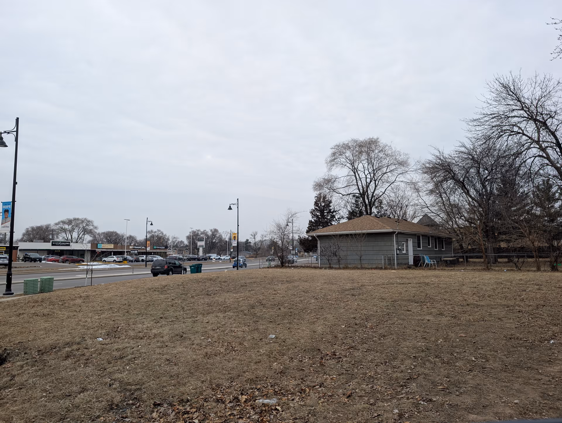 A small single-story house on a grassy lot beside a busy street with shops and cars under an overcast sky.