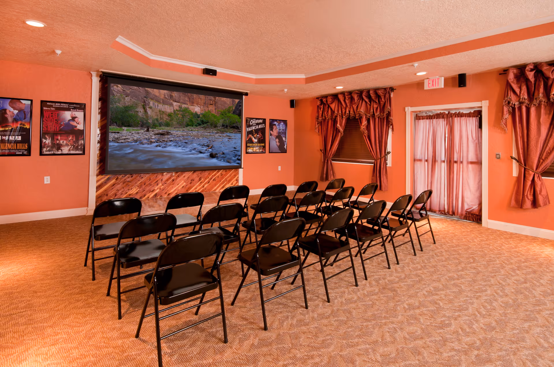 A small theater or presentation room with rows of black folding chairs facing a large screen displaying a nature scene of a river and cliffs. The walls are painted orange and decorated with movie posters. There are red curtains on the windows and a door with a curtain. The carpet is patterned in shades of orange.