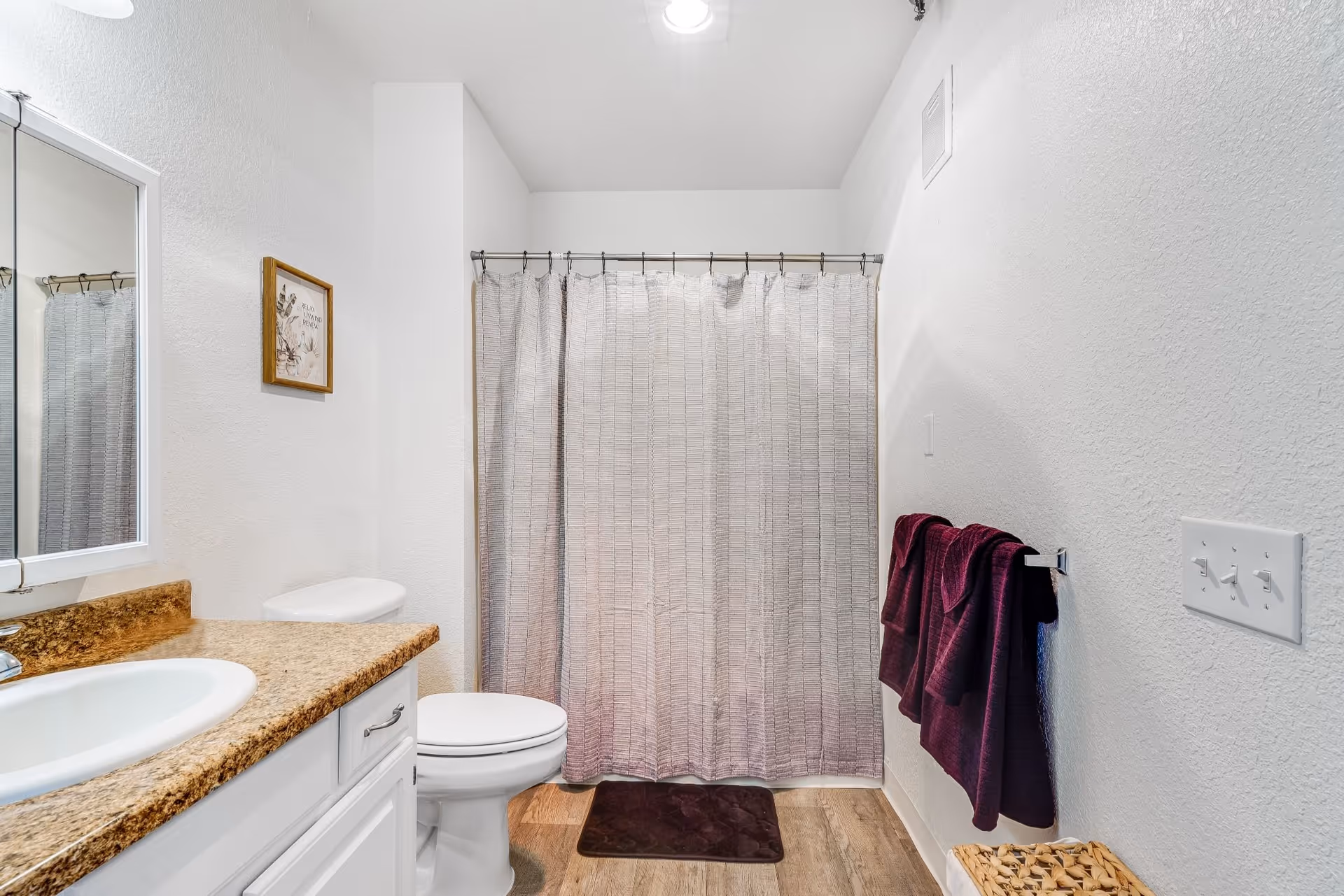Bright bathroom with a sink and countertop, toilet, shower with a curtain, and purple towels on a wall-mounted rack.