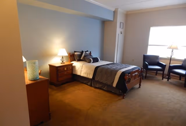 Neatly furnished bedroom with a single bed, nightstands and lamps, two chairs by a window, and carpeted floor.