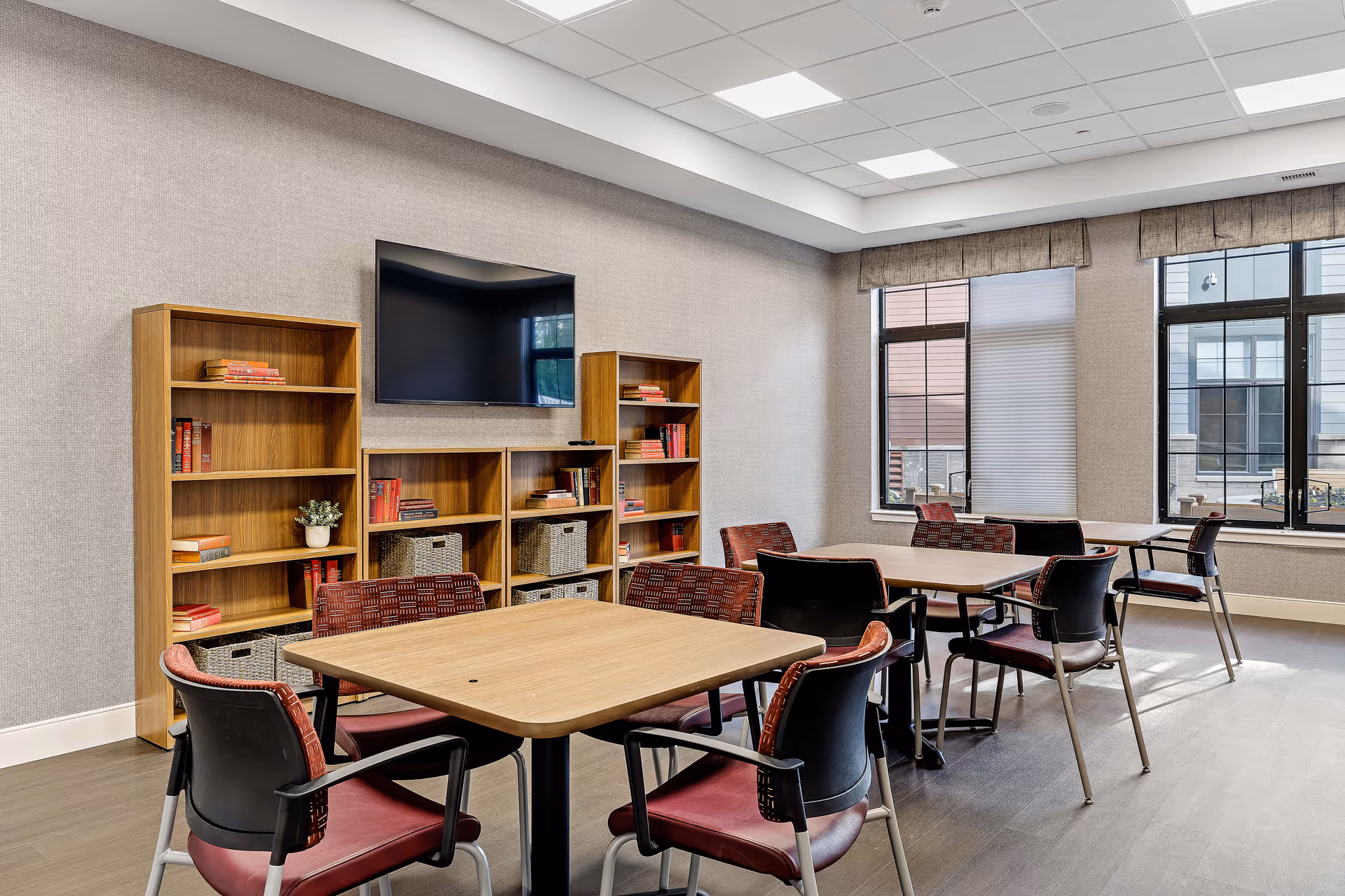 Bright communal activity/dining room with several tables and chairs, bookshelves, a wall-mounted TV, and large windows.