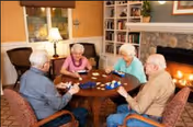 Four elderly people sitting around a round wooden table playing a card game in a cozy living room with a fireplace, bookshelves, and a lamp.