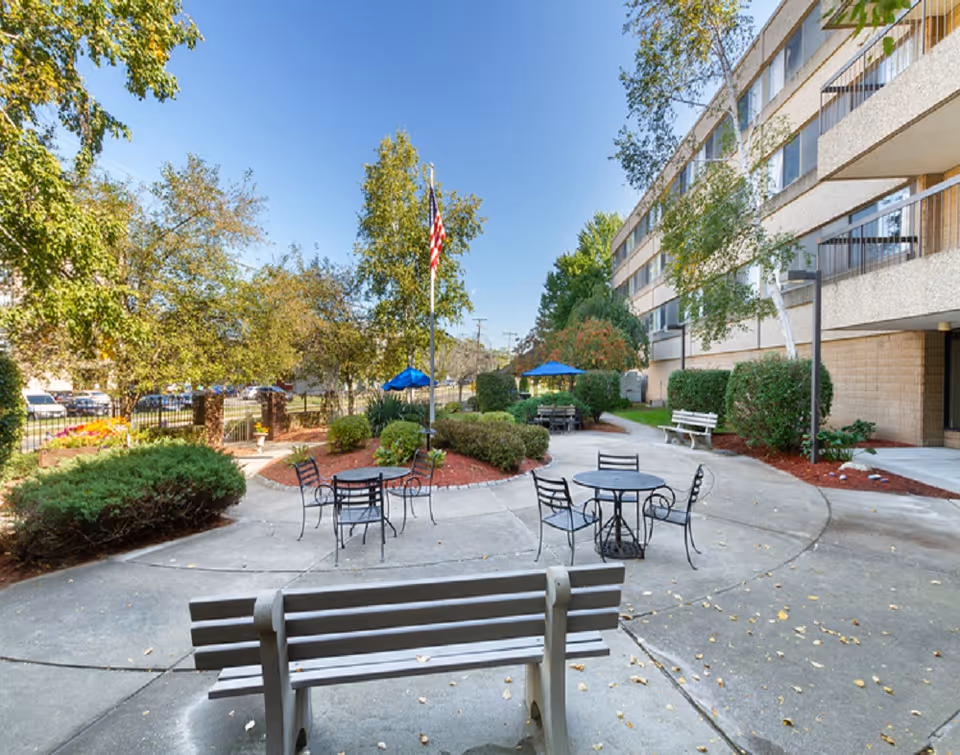 Outdoor courtyard with benches, round metal tables and chairs, landscaped shrubs, an American flag, and a multi-story building facade.