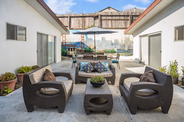 Outdoor patio area with wicker-style seating including two armchairs, a loveseat with blue and patterned cushions, and a coffee table with a plant. In the background, there is a dining table with chairs under a large umbrella. The area is surrounded by white buildings with sliding glass doors and potted plants. A mural of the San Francisco skyline, Golden Gate Bridge, and a cable car is painted on a wooden fence.
