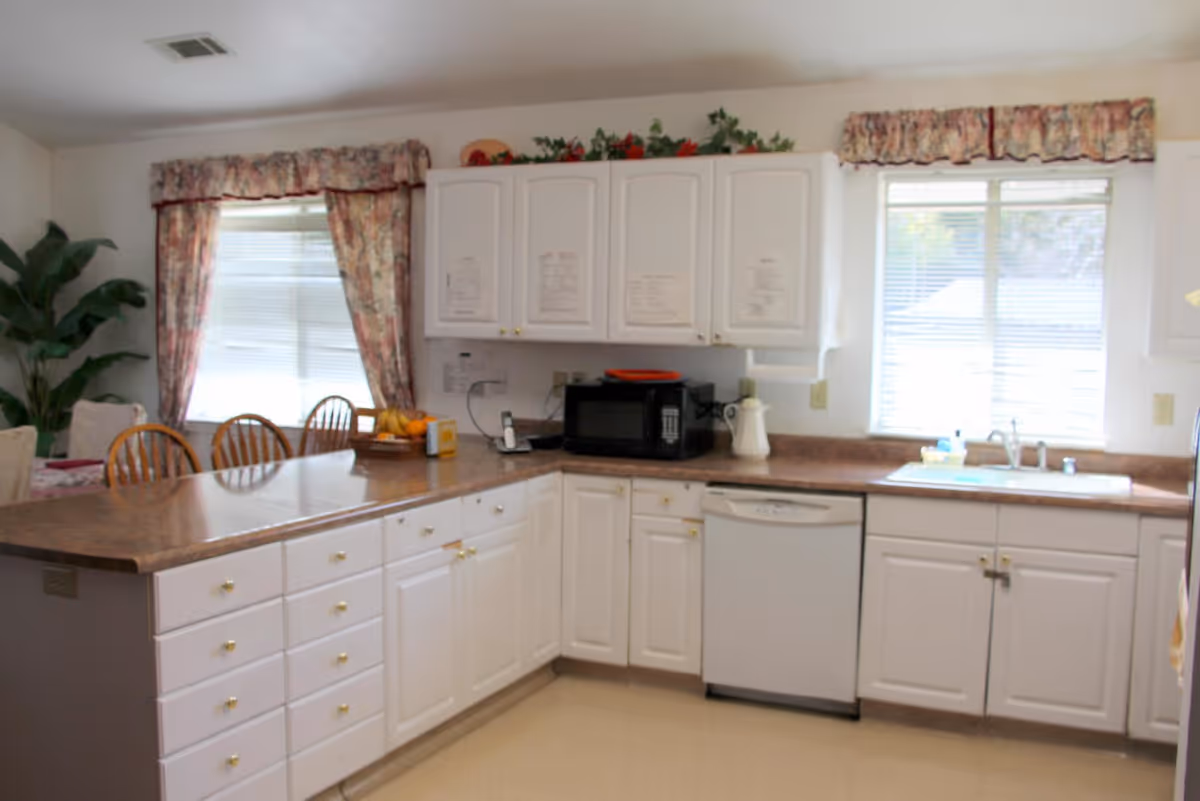 A bright kitchen with white cabinets and drawers, a brown countertop, a black microwave, a white dishwasher, and a white sink under a window with blinds. There are floral curtains on the windows and a green plant in the corner. A dining area with wooden chairs is partially visible.