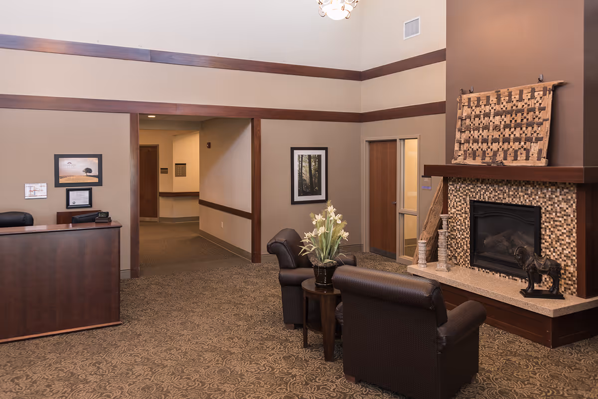 A senior living facility lobby with two leather chairs, a small table with flowers, a fireplace, and a reception desk.