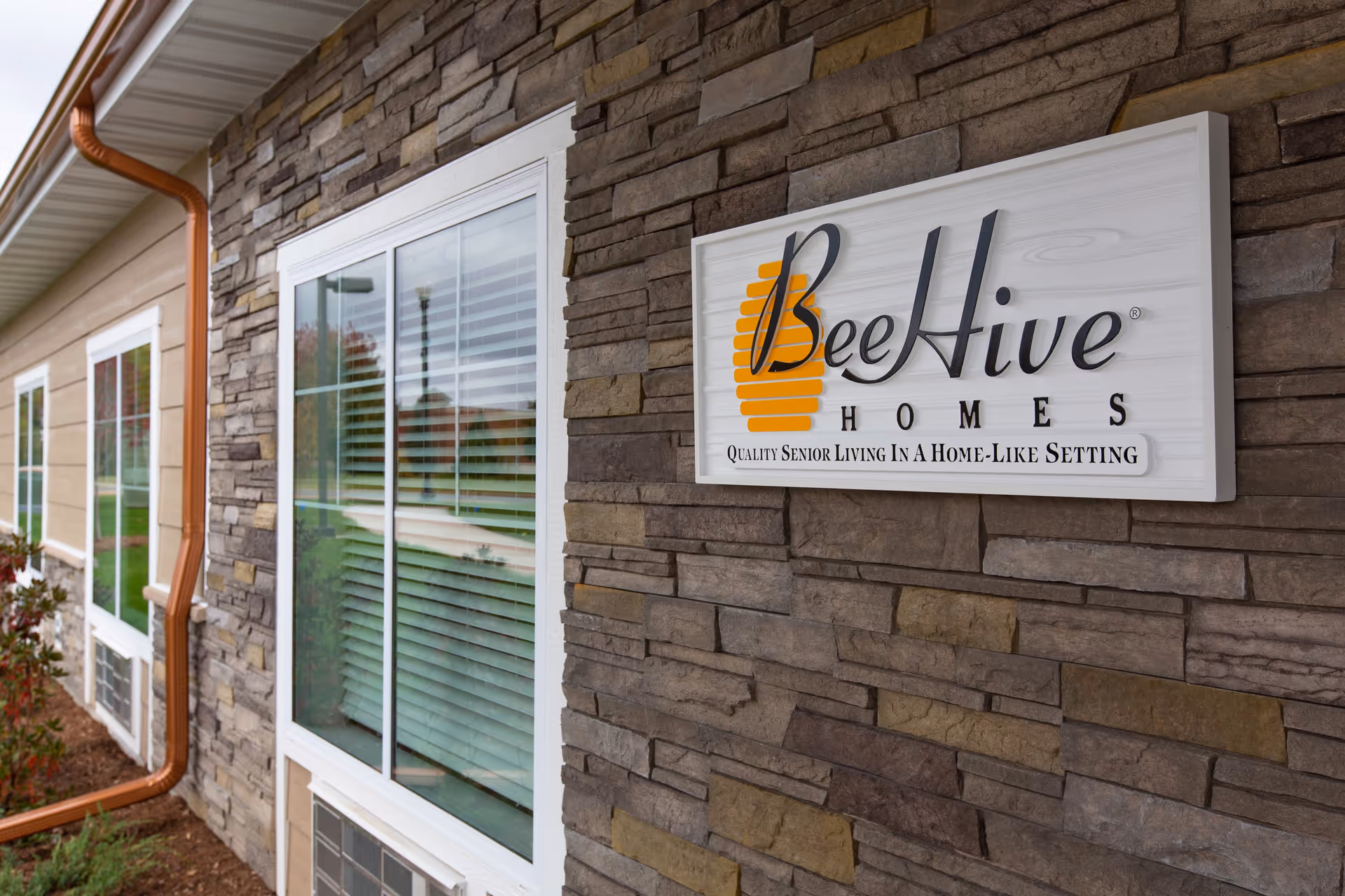Exterior wall of a building with stone veneer and beige siding, featuring multiple white-framed windows with blinds. A white sign with the BeeHive Homes logo and the tagline 'Quality Senior Living In A Home-Like Setting' is mounted on the stone wall.