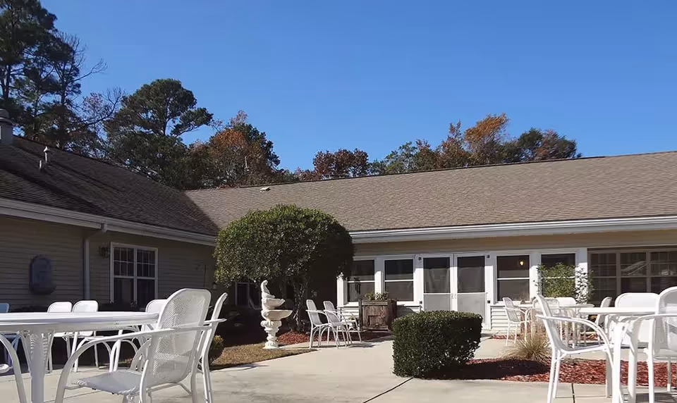 Outdoor patio area at Savannah Cottage of Lakeland with white metal tables and chairs, trimmed bushes, a decorative fountain, and a building with multiple windows and doors under a clear blue sky.