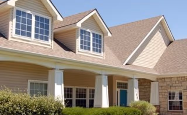 Exterior view of a residential-style building with beige siding, white columns, multiple windows, and a brown shingled roof under a clear blue sky. There are green bushes in front of the building.