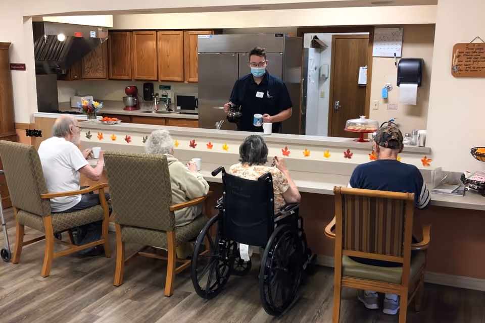 Four elderly individuals seated at a counter in a senior living facility, with one person in a wheelchair. A staff member wearing a mask is behind the counter pouring coffee. The kitchen area behind the counter has wooden cabinets, a refrigerator, and various kitchen appliances. The counter is decorated with small colorful leaf cutouts.