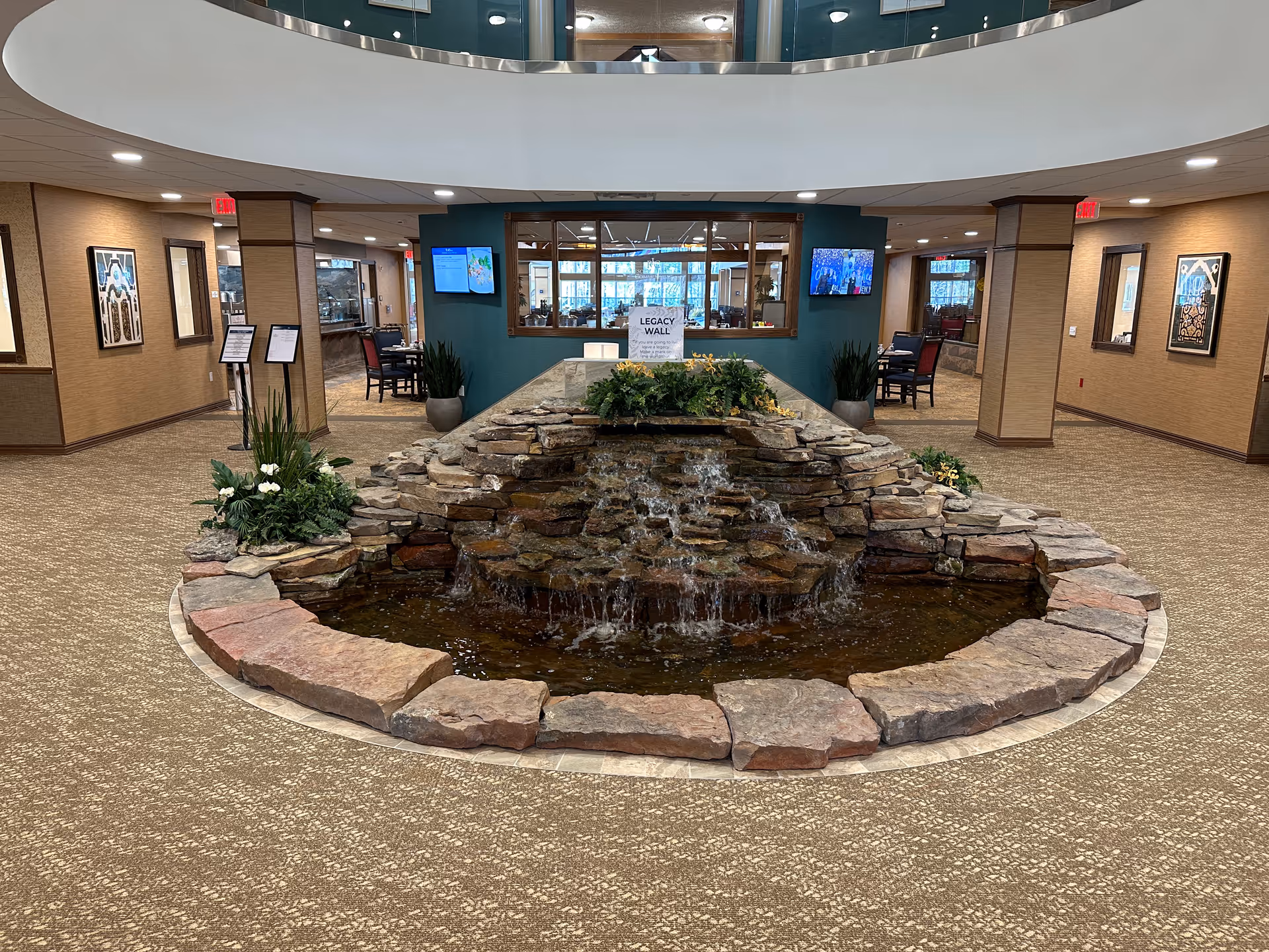 Indoor lobby with a central tiered rock water fountain surrounded by carpeting, plants, and seating areas.