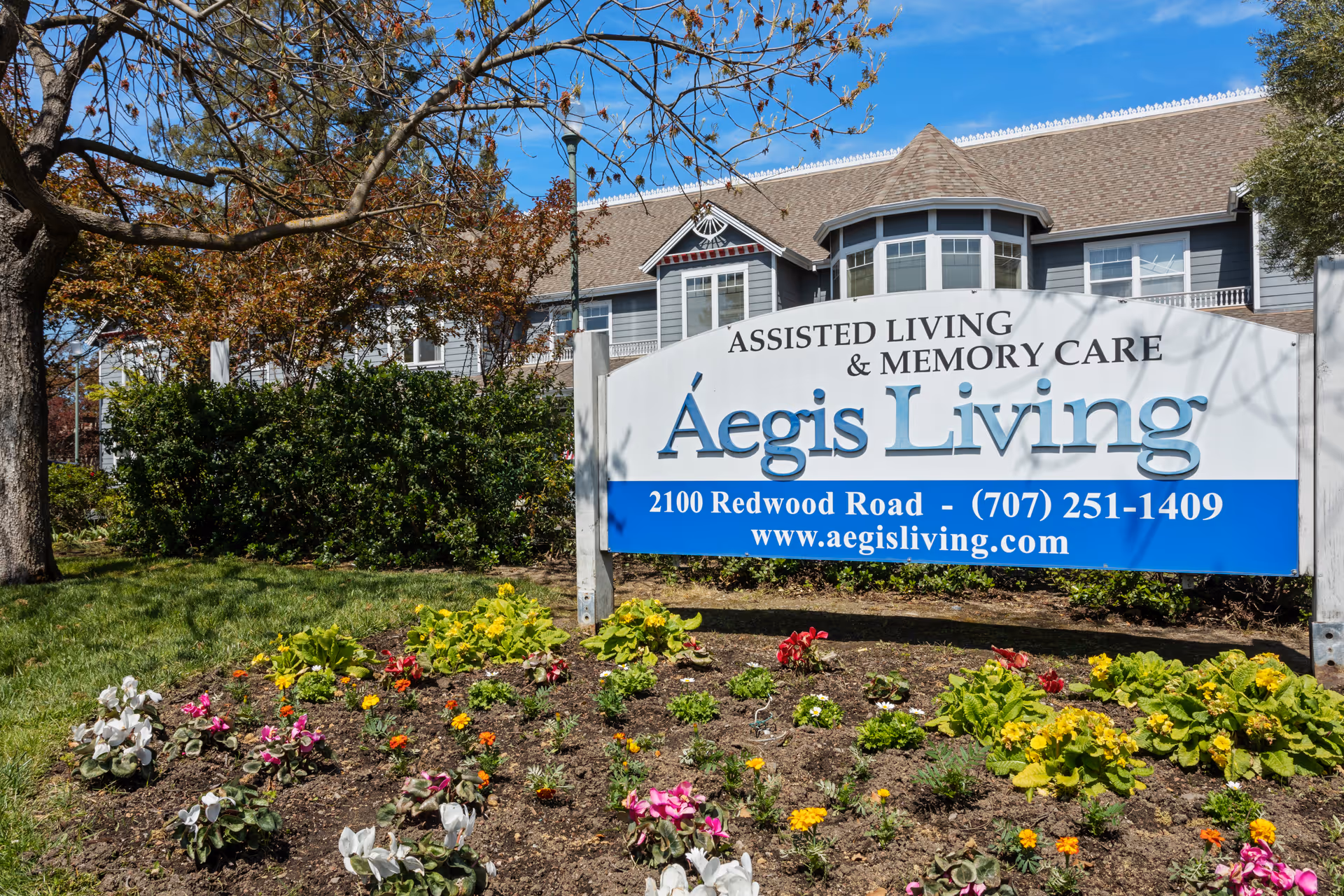 Entrance sign for Aegis Living assisted living & memory care with a flowerbed and the facility building behind it.