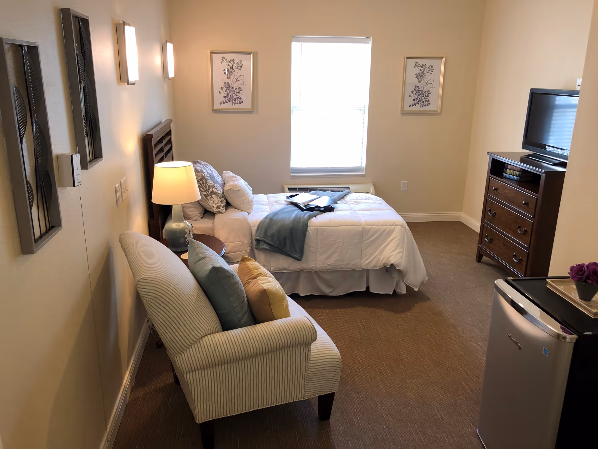 A cozy bedroom in a senior living facility featuring a single bed with white bedding and decorative pillows, a small armchair with cushions, a wooden dresser with a flat-screen TV on top, a mini refrigerator, and wall decorations including framed floral prints and metal leaf art. The room is softly lit by wall sconces and a table lamp.