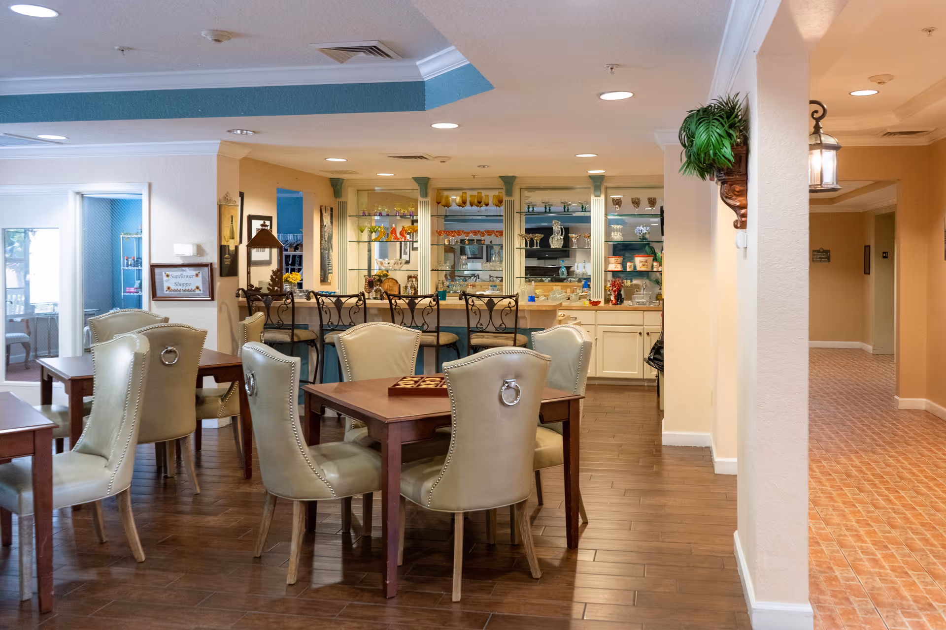 Interior view of a senior living facility common area with several tables and cushioned chairs arranged for socializing or games. In the background, there is a counter with bar stools and shelves displaying decorative glassware and collectibles. The space has warm lighting, wooden flooring, and beige walls with some green accents.