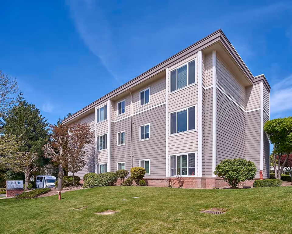 Three-story beige and taupe senior living building with multiple windows, landscaped lawn, shrubs and clear blue sky.