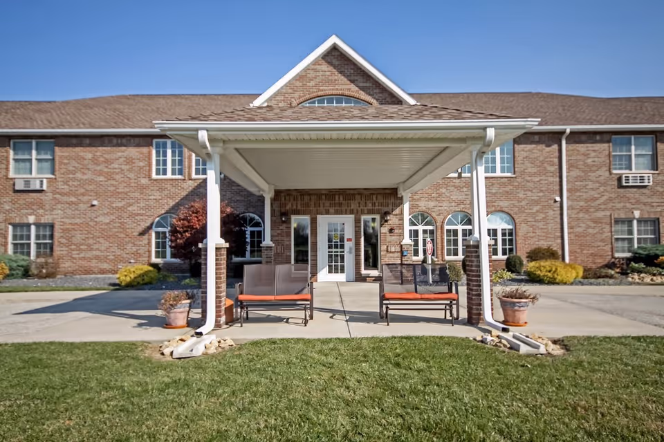 Front exterior view of a two-story brick building with a covered entrance supported by white columns. There are two benches with orange cushions under the entrance canopy, potted plants on either side, and a well-maintained lawn in the foreground. The building has multiple windows, some with air conditioning units.