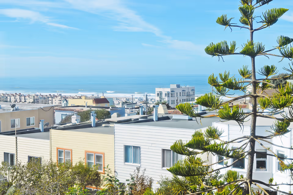 View of residential buildings with a tree in the foreground and the ocean in the background under a clear blue sky.