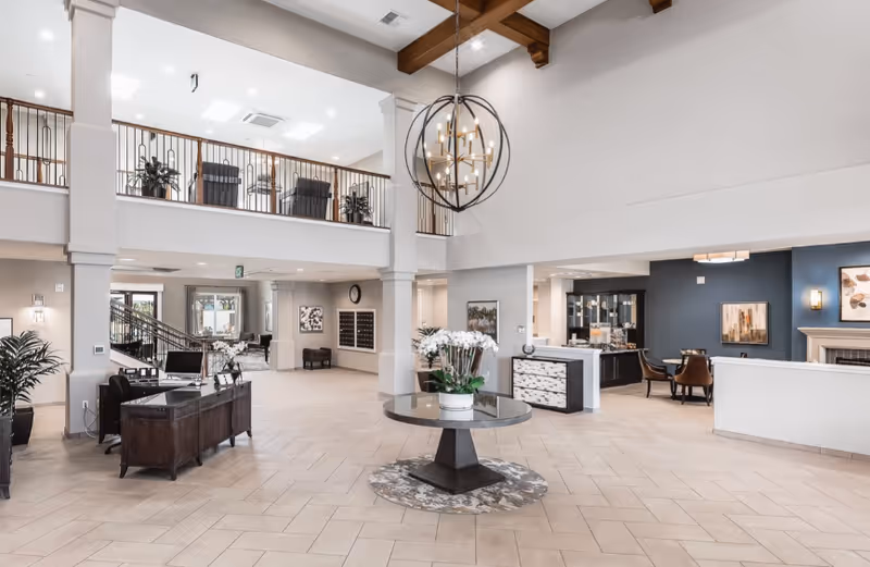 Spacious and well-lit senior living facility lobby with high ceilings and exposed wooden beams. A round table with a potted white orchid is centered on a circular rug. To the left, a dark wooden reception desk with computer monitors is visible. The upper floor has a railing with plants and seating. The right side features a seating area with chairs, a fireplace, and framed artwork on the walls.
