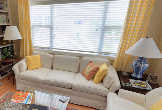 Bright living room with a beige sofa and colorful throw pillows in front of a large window with yellow-check curtains, flanked by lamps and a coffee table.