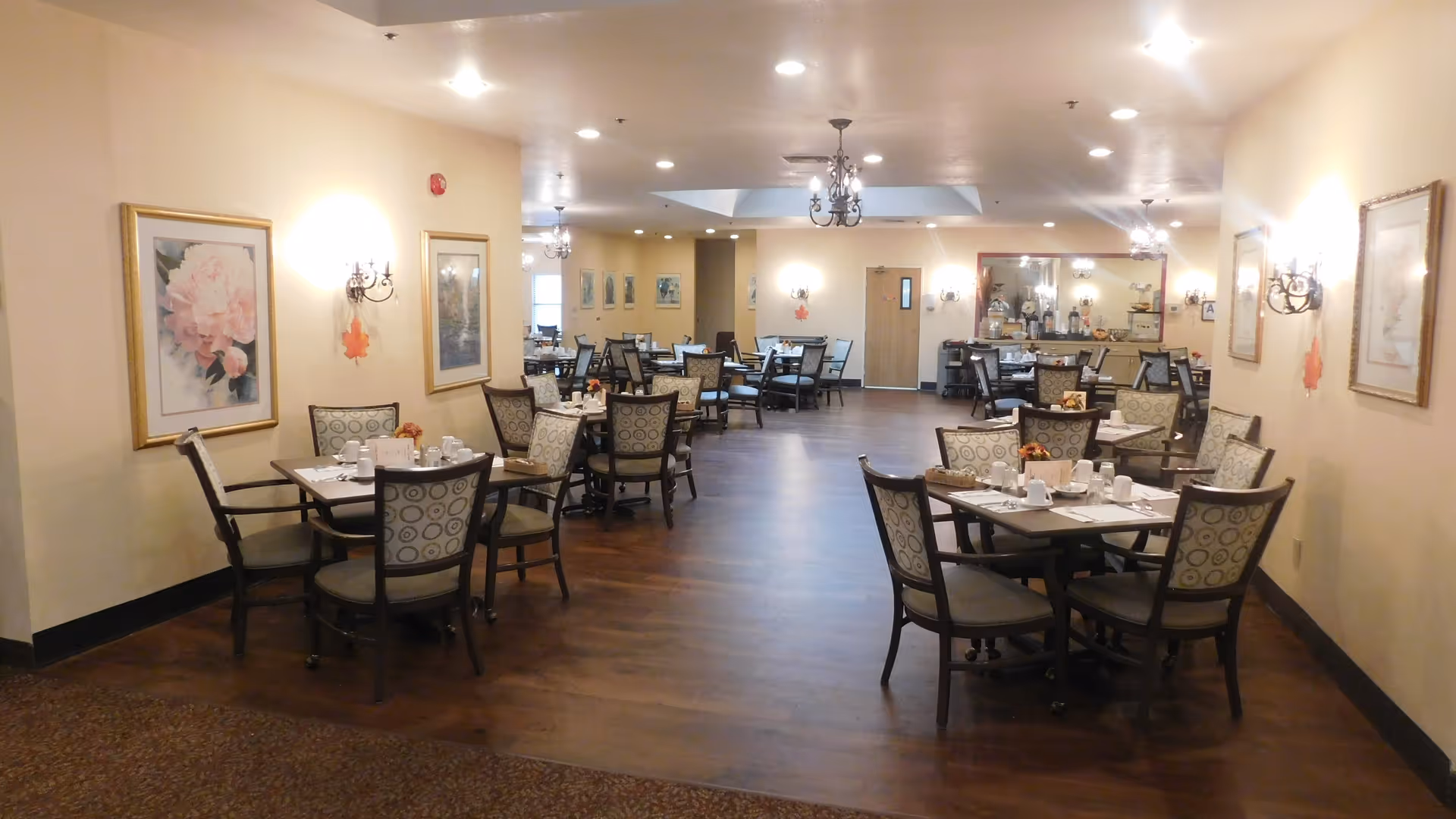 Large dining room with multiple set tables and upholstered chairs, chandeliers, and framed artwork on the walls.