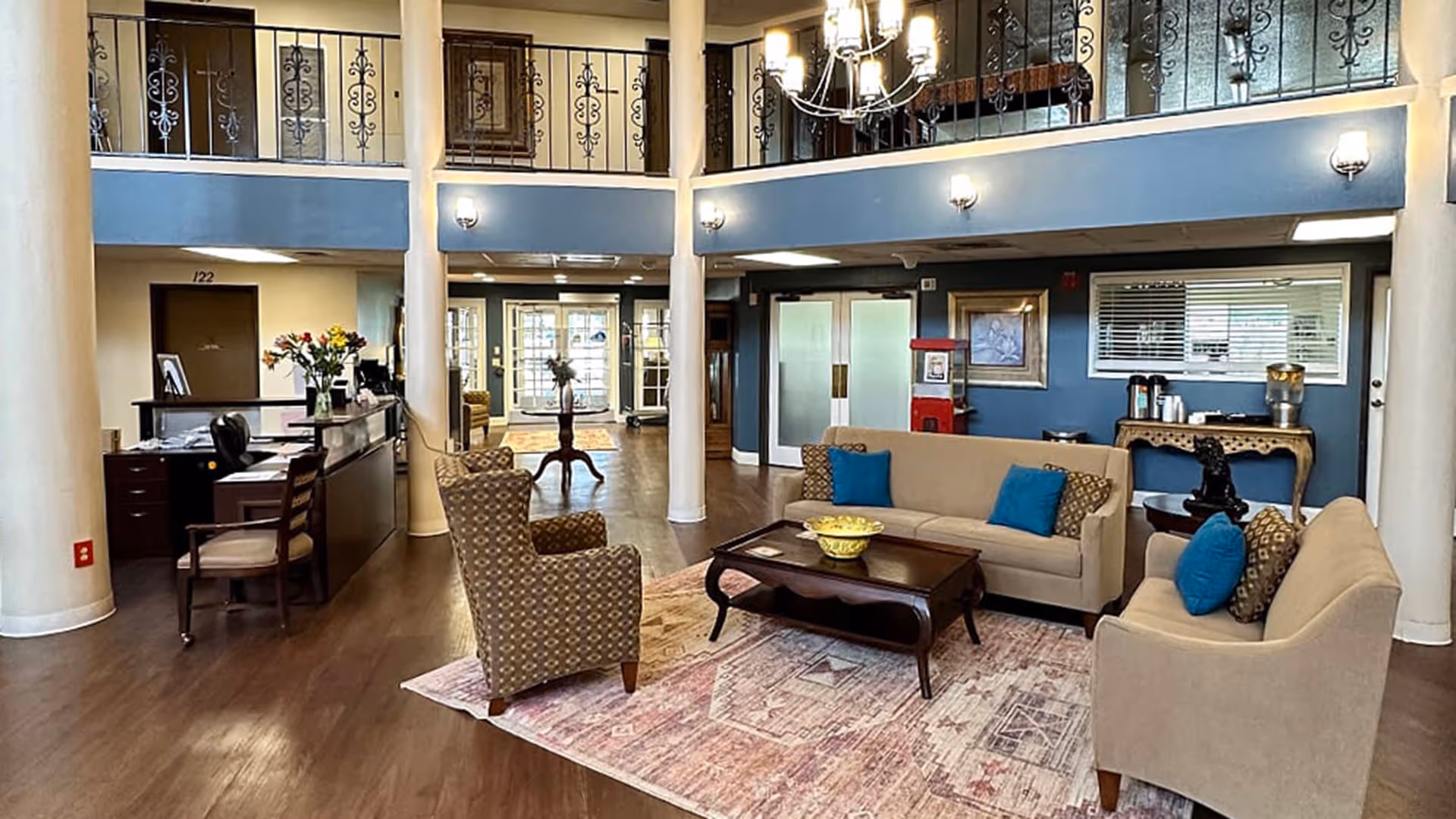 A spacious senior living facility common area with wooden floors, beige sofas with blue and patterned cushions, a patterned armchair, and a wooden coffee table on a large area rug. There is a reception desk with a chair and flowers on the left side, and a small table with coffee and water dispensers on the right. The room features white columns, blue walls with decorative railings on the upper level, and multiple light fixtures including wall sconces and a chandelier. Double glass doors and windows are visible in the background.