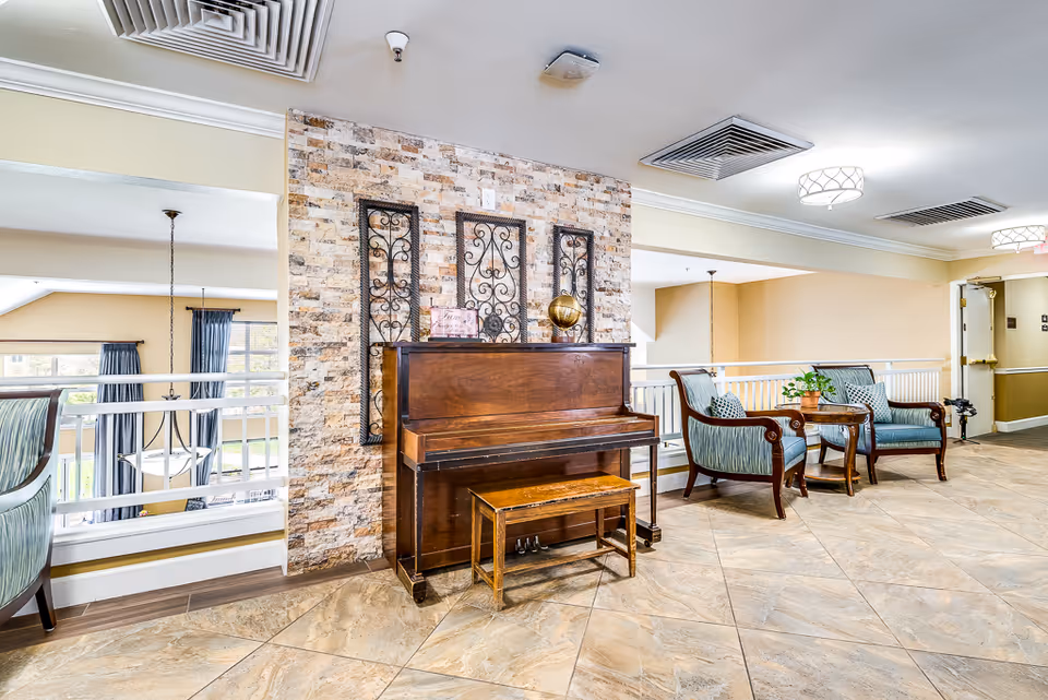 A bright and spacious senior living facility common area featuring a wooden upright piano with a matching bench against a stone accent wall. Next to the piano are two upholstered armchairs with a small wooden table between them, decorated with a potted plant. The area has tiled flooring, light-colored walls, and ceiling lights, with a railing overlooking a lower level with large windows and curtains.