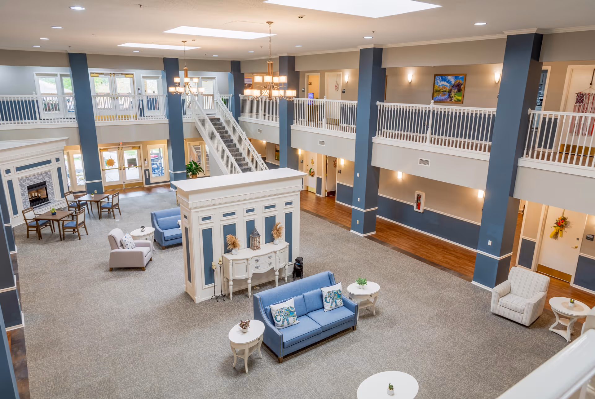 Spacious two-story atrium with seating areas, a central staircase, and decorative furnishings in a senior living facility.