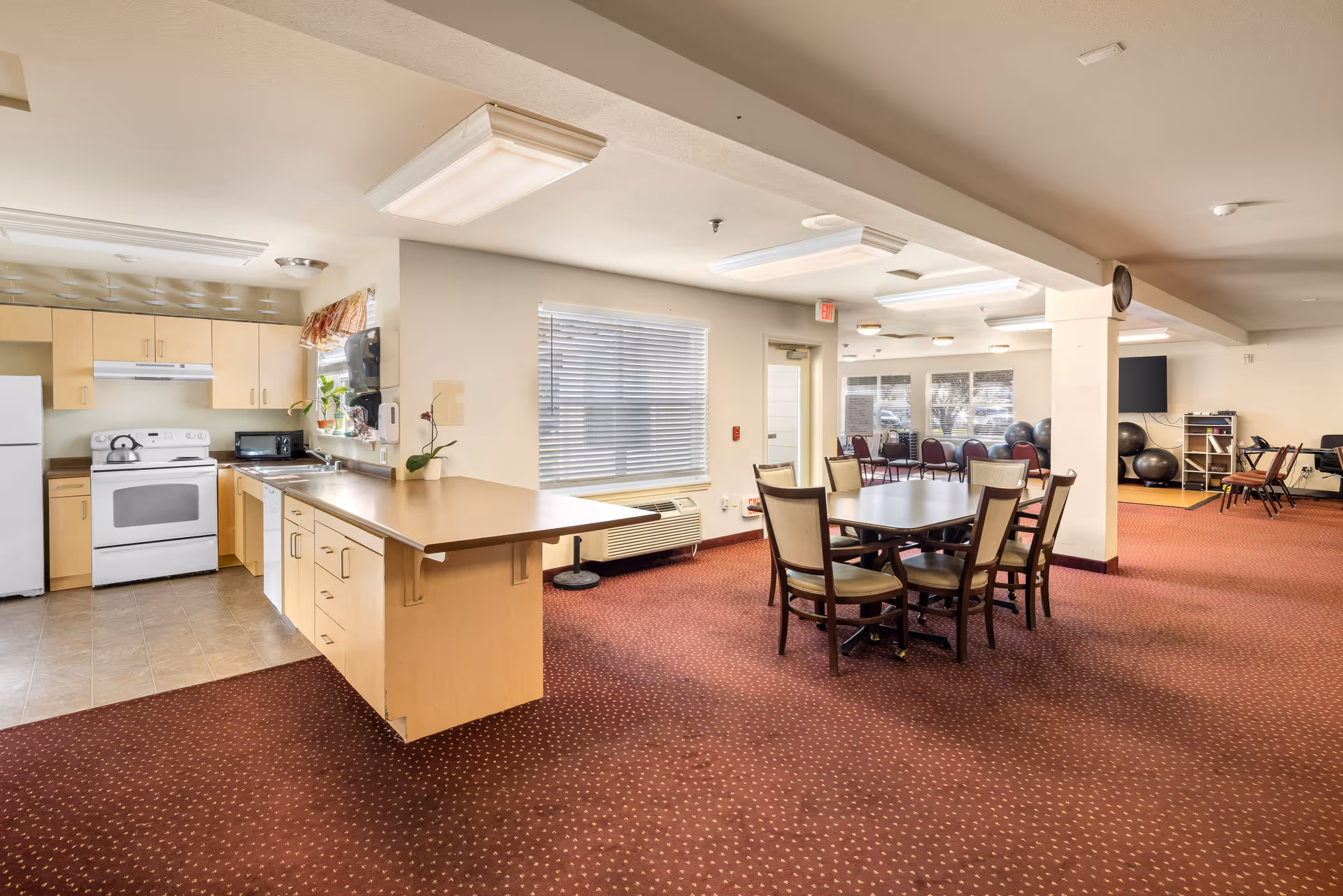 Interior view of a senior living facility showing a kitchen area with light wood cabinets, white appliances including a refrigerator and stove, and a countertop extending into a dining area with a table and chairs. The room has carpeted flooring, large windows with blinds, and fluorescent ceiling lights. In the background, there are additional chairs and exercise balls near a wall-mounted TV.