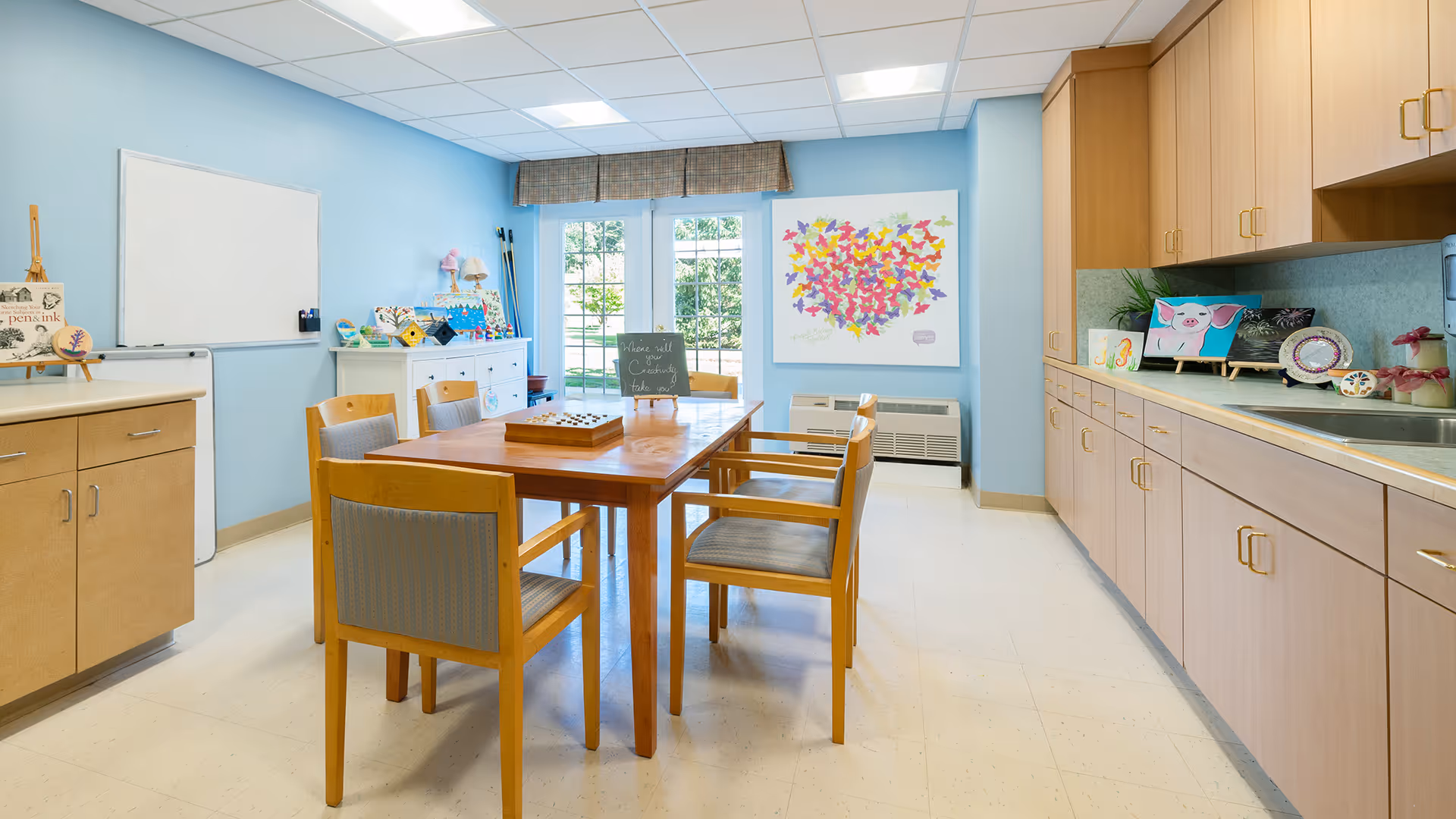 A bright activity room with light blue walls, a wooden table surrounded by six chairs, cabinets with art supplies and decorations, a whiteboard on the wall, and a large window with a view of greenery outside.