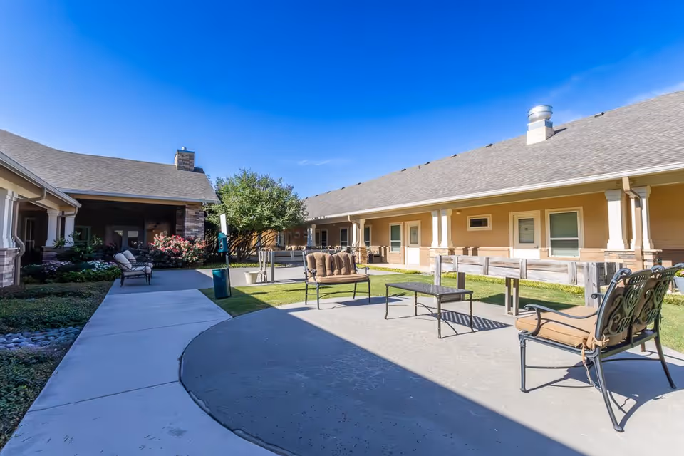 Outdoor courtyard area of an assisted living facility with cushioned metal chairs and benches arranged on a concrete patio. The courtyard is surrounded by single-story buildings with beige walls and white trim under a clear blue sky. There are some bushes and a tree near the buildings, and a paved walkway runs through the courtyard.