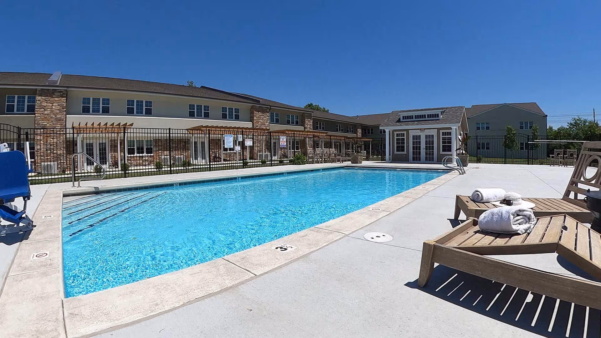 Outdoor swimming pool at Sycamore Reserve Senior Living with clear blue water, surrounded by a concrete deck. Wooden lounge chairs with rolled white towels are placed on the right side. The pool area is fenced, and behind it is a two-story building with stone and beige siding under a clear blue sky.