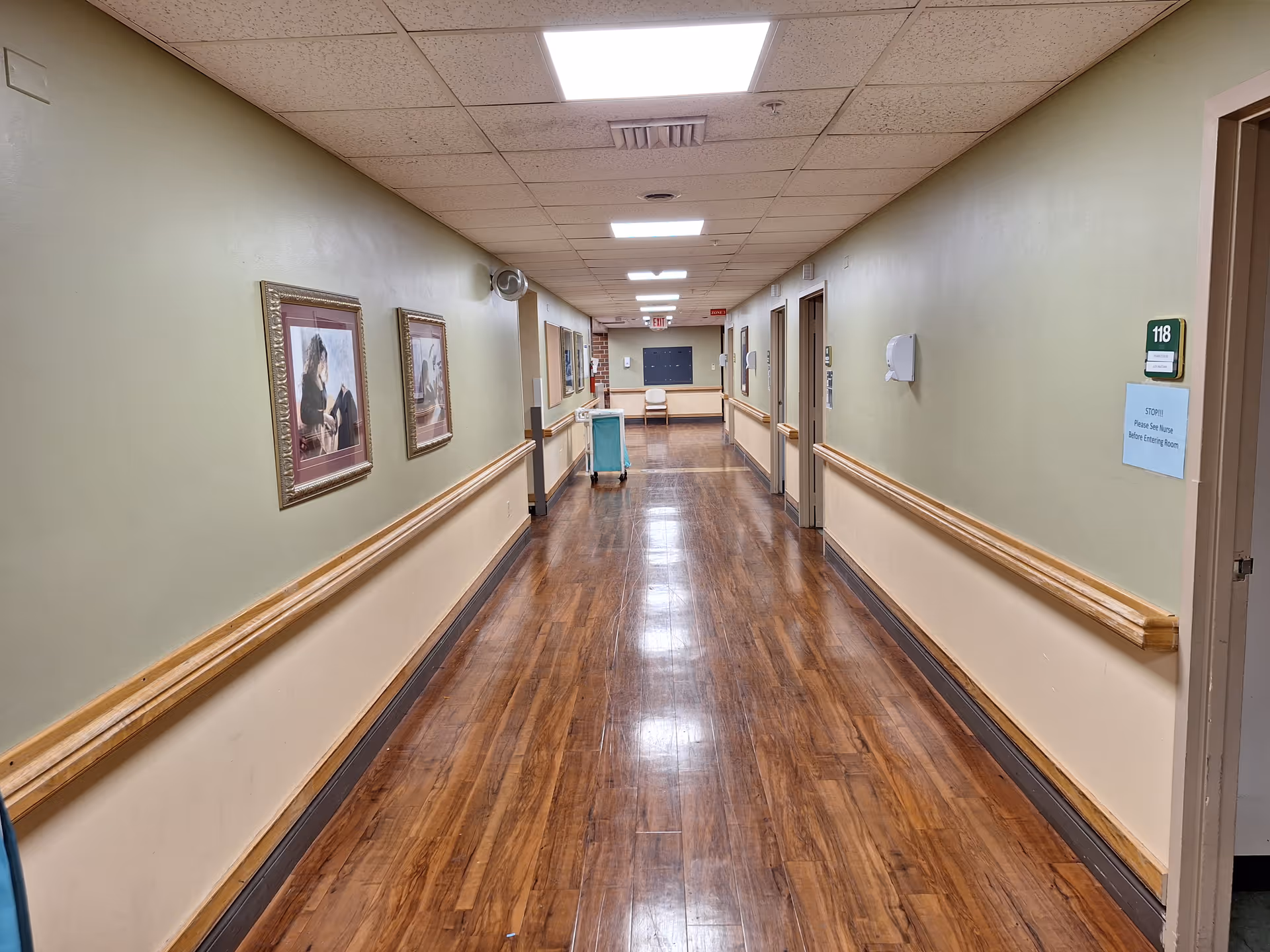 A long, clean hallway in a senior living facility with wooden floors, beige walls, and handrails on both sides. Several framed pictures hang on the left wall, and multiple doors line the right side. A green room number sign labeled 118 is visible on the right, along with a blue notice. The ceiling has recessed lighting panels.