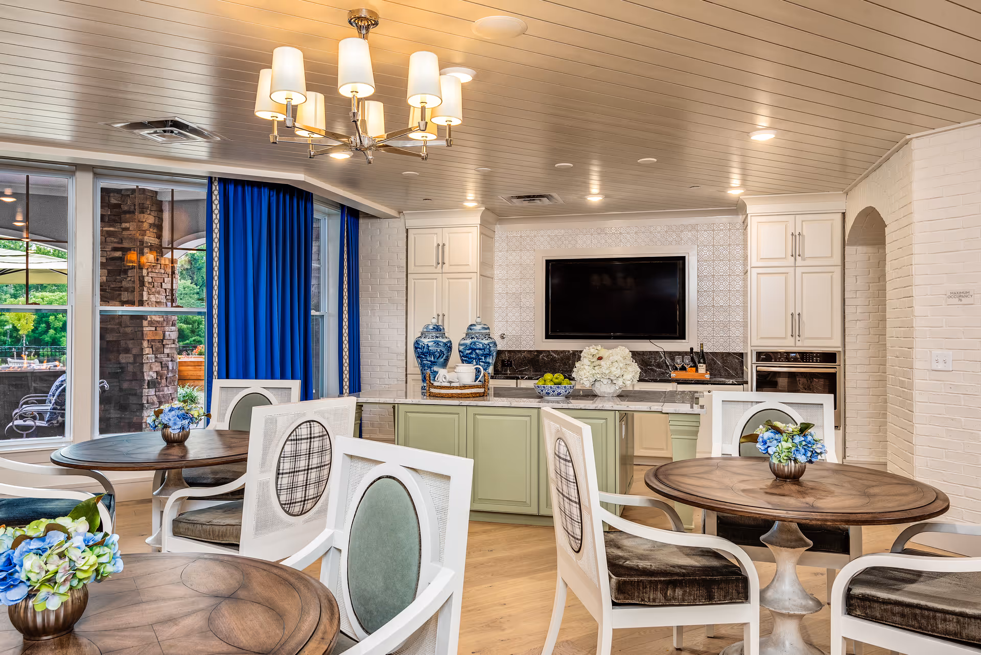 A bright and elegant common area with round wooden tables and white chairs featuring cushioned seats and backs. The room has a light wood floor, white paneled ceiling with recessed lighting, and a chandelier. There is a kitchen area with green cabinets, a black countertop, decorative blue and white vases, and a mounted flat-screen TV on a tiled wall. Large windows with blue curtains show an outdoor patio area with stone pillars and greenery.