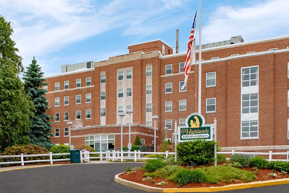 Exterior view of a multi-story brick building with numerous windows, surrounded by greenery and a curved driveway. In front of the building is a sign that reads 'The Highlands' with the tagline 'Life Care Centers of America' and an American flag on a flagpole.