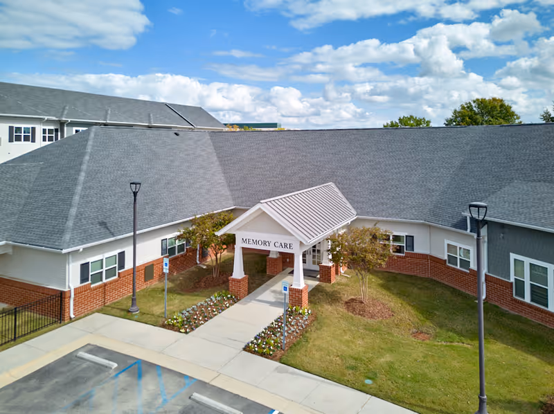 Exterior view of a single-story building with a covered entrance labeled 'Memory Care'. The building has a gray shingled roof, white siding with red brick accents, and landscaped areas with small trees and flowers. There are two lamp posts and a parking area with handicap parking spaces in front.