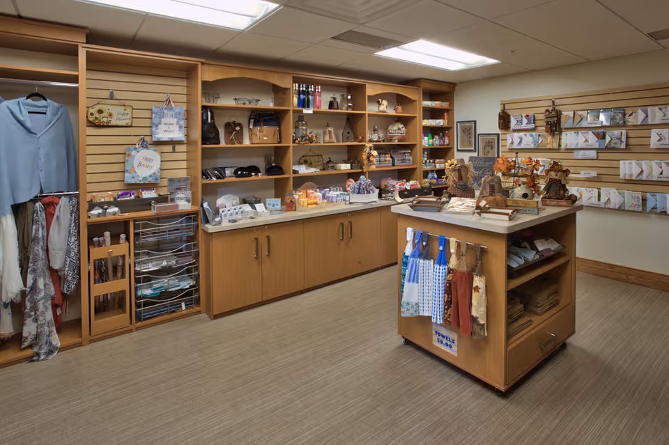 Small interior gift shop with wooden shelving and a central display island holding towels, clothing, and assorted gifts.