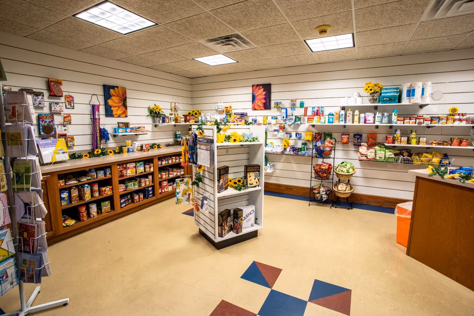 Interior view of a small convenience store or shop area inside a senior living facility, with shelves stocked with various food items, snacks, and personal care products. There is a display rack with greeting cards on the left and a central shelving unit with books and other items. The walls have sunflower decorations and paintings, and the floor has a patterned tile design.