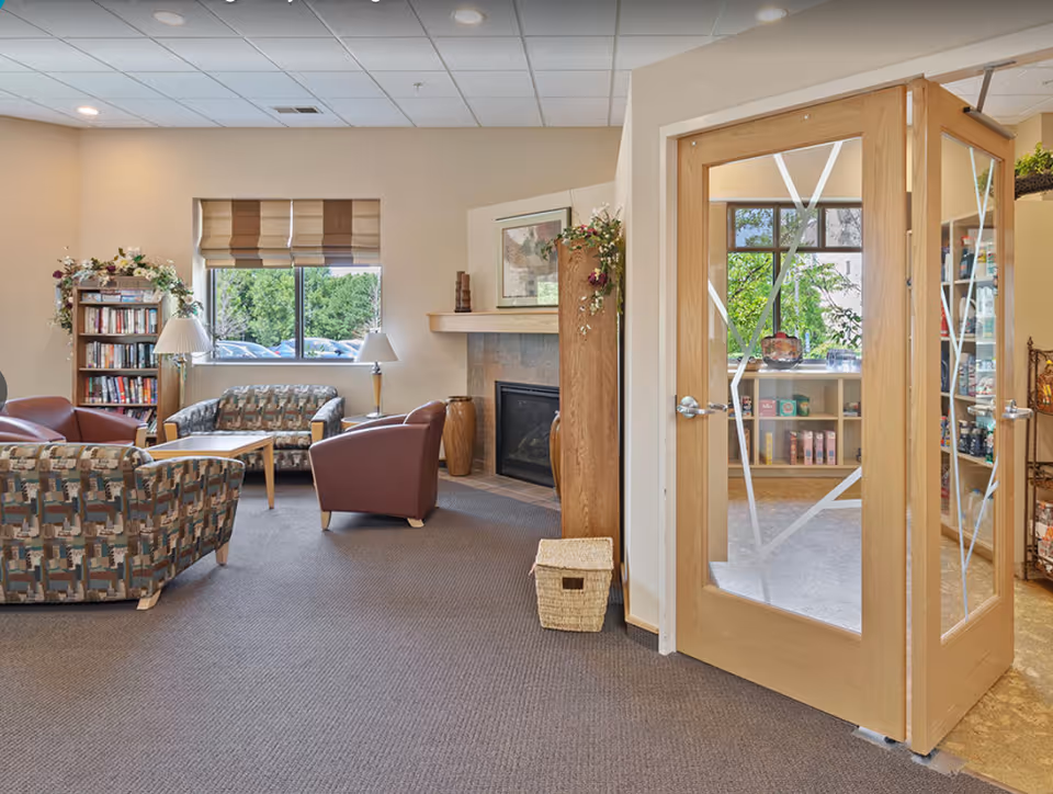 A senior living lounge with upholstered chairs and sofas around a coffee table, a fireplace, bookshelf, and glass doors leading to another room.