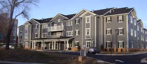 Three-story gray Symphony Manor senior living building with a covered entrance and parked car.