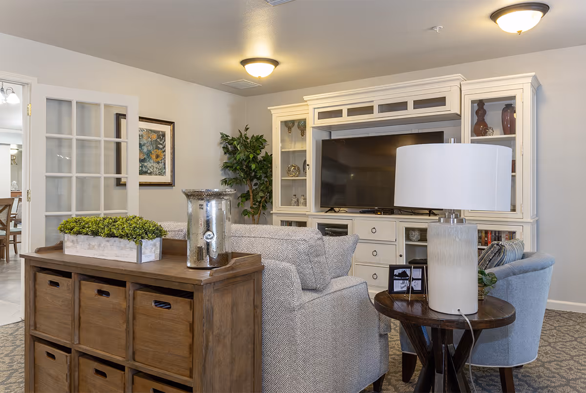 A cozy living room area with a large white entertainment center holding a flat-screen TV. In front of the TV, there are two upholstered chairs and a wooden side table with a white lamp. A wooden storage unit with baskets and a decorative plant is also visible. The room has neutral walls, carpeted flooring, and ceiling lights. A framed floral picture and a potted plant are seen near a glass-paneled door leading to another room.