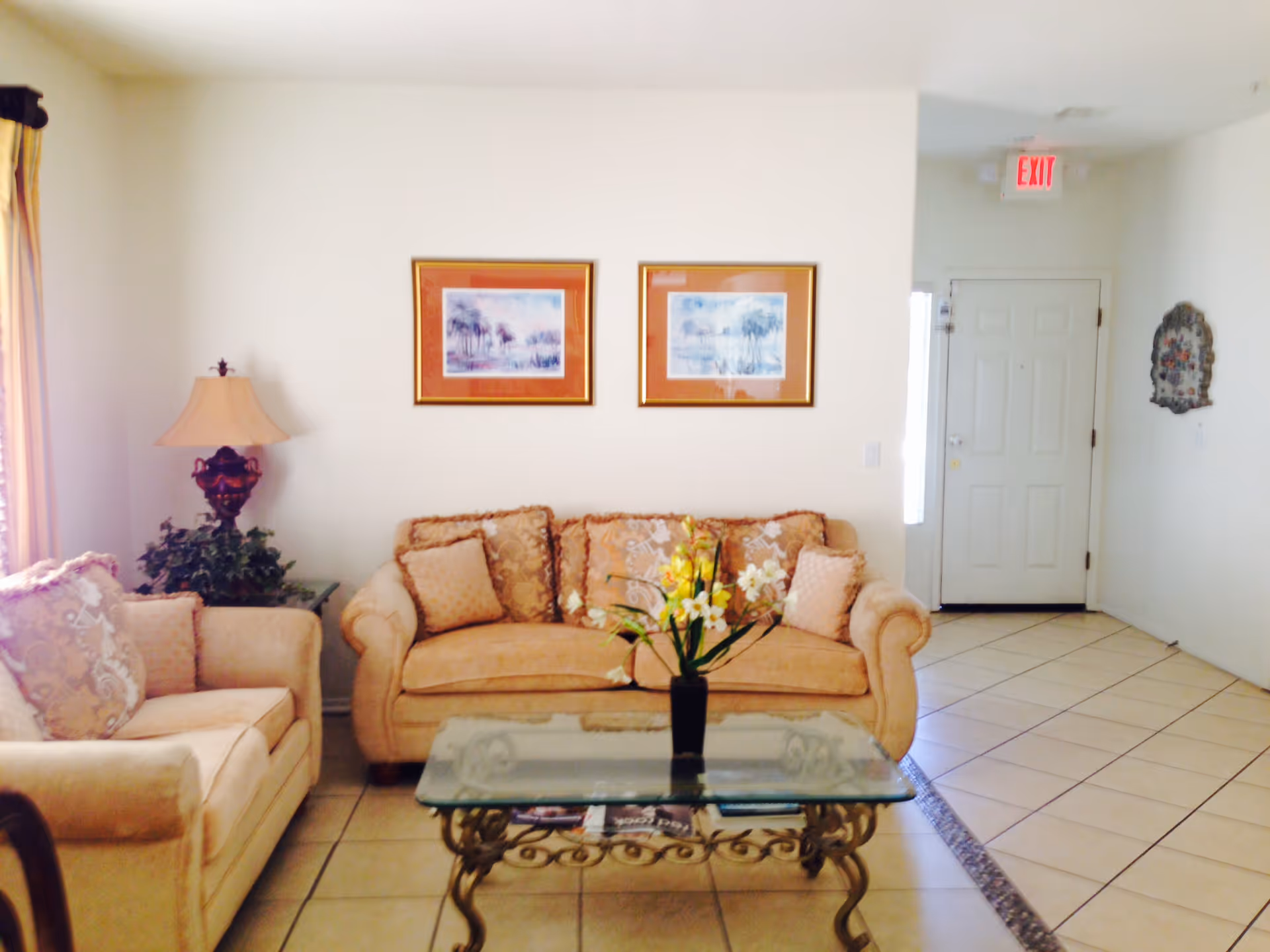 A cozy living room with beige upholstered sofa and armchair, a glass coffee table with a floral arrangement, two framed pictures on the wall, a table lamp, and a white front door with an exit sign above it.