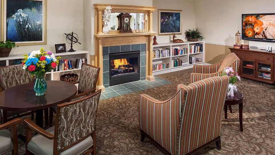 A cozy living room area with a lit fireplace surrounded by a wooden mantel. There are two striped armchairs facing the fireplace, a round table with a vase of colorful flowers, bookshelves filled with books, and a TV mounted on a wooden cabinet displaying an image of orange flowers. The room is decorated with paintings, plants, and a patterned carpet.
