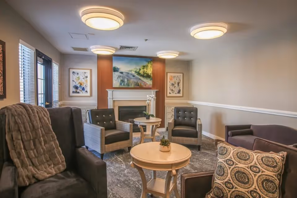 A cozy senior living facility lounge with comfortable armchairs and a sofa arranged around two round wooden tables. The room features a fireplace with a landscape painting above it and two floral artworks on either side. The walls are painted beige with white trim, and the ceiling has modern circular light fixtures. A window with blinds and a door are visible on the left side.
